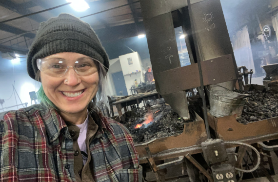 Cindy smiles at the camera posing in front of her blacksmithing equipment.