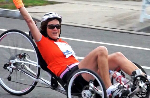Tracie on a trike smiling at the camera and waving one arm in the air while passing the camera